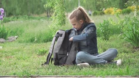 a girl, a primary school student, sits on the green grass in the park. she is engrossed in browsing her smartphone, her school backpack is nearby.