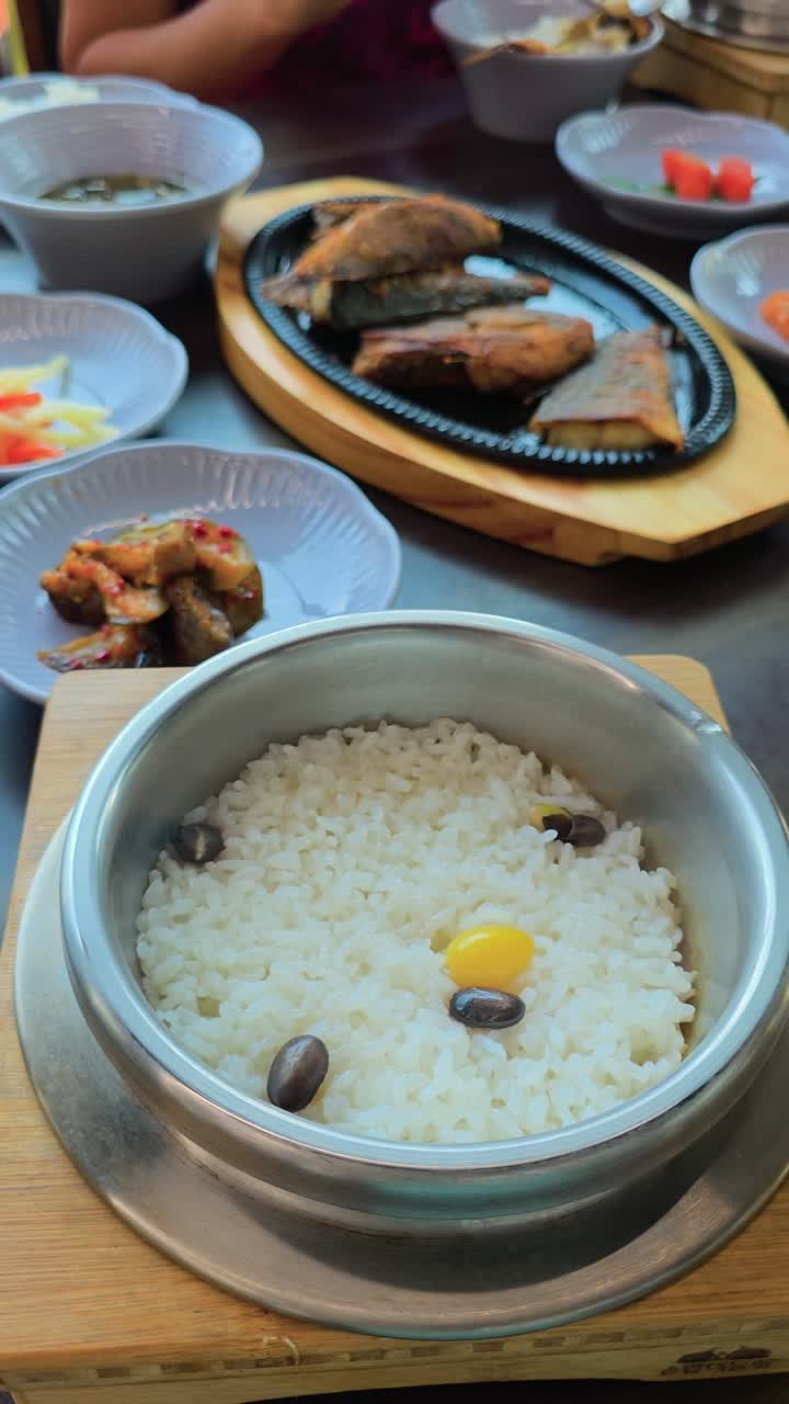 A man's hand opens a steaming hot metal pot to reveal freshly cooked rice with beans and chestnut, served in a Korean traditional restaurant with assorted side dishes and fried fish - vertical