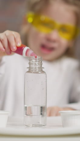 joyful little girl with protective goggles drips red liquid carefully with asian and african-american teachers at fun science lesson closeup