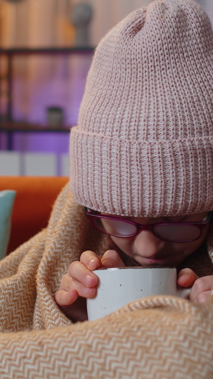 Ragazza malata che indossa un cappello avvolto in un quadri si siede da sola tremando di freddo sul divano bevendo tè caldo