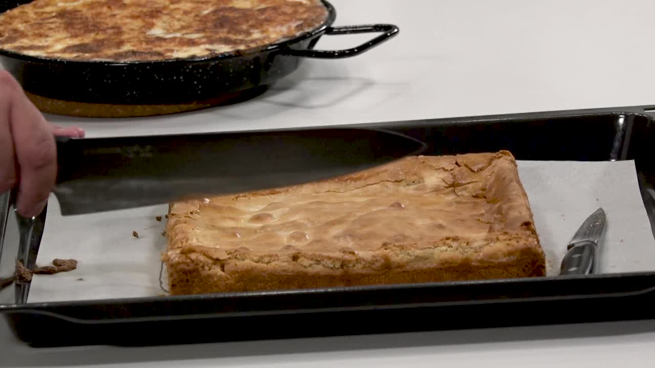 A baker skillfully slices into a freshly baked dessert on a baking tray. The cake appears golden brown and fluffy, ready to be served. Delicious aromas fill the air, tempting everyone nearby