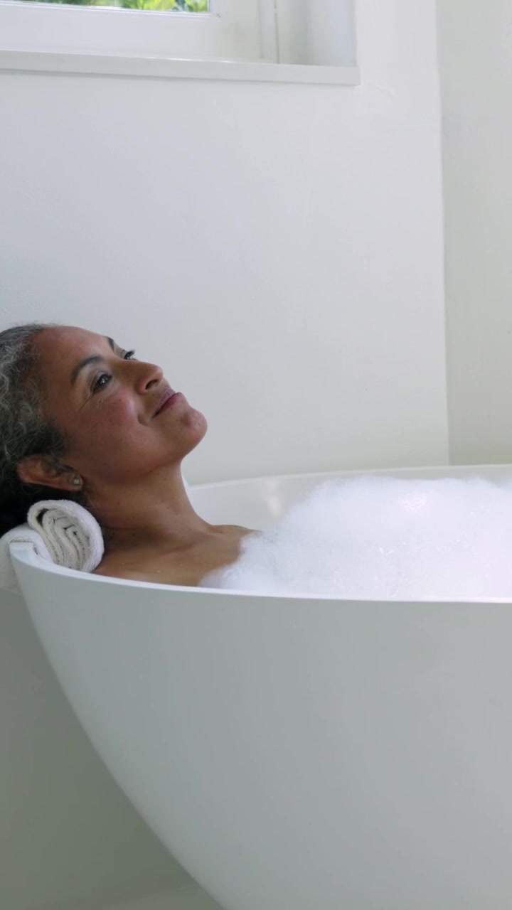 Video verticale: Anziana donna africana che si muove, sollevando le ginocchia e spostando la schiuma nella vasca da bagno a casa, rilassandosi.