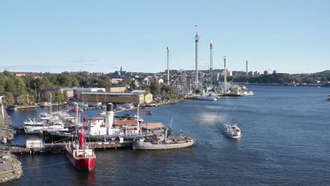 Boats-in-dock-and-cruising-by-with-Gröna-Lund-in-background-during-sunny-evening-in-Djurgården,-Stockholm,-Sweden