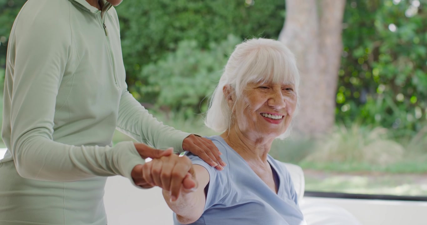 African woman smiling, guiding senior woman using hands, stretching arm from chair for mobility
