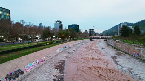 Dolly-De-Drones-En-Una-Toma-Del-Agua-Del-Río-Mapocho-Que-Fluye-A-Través-De-Santiago-De-Chile-Con-Gran-Fuerza-En-Una-Puesta-De-Sol-Nublada