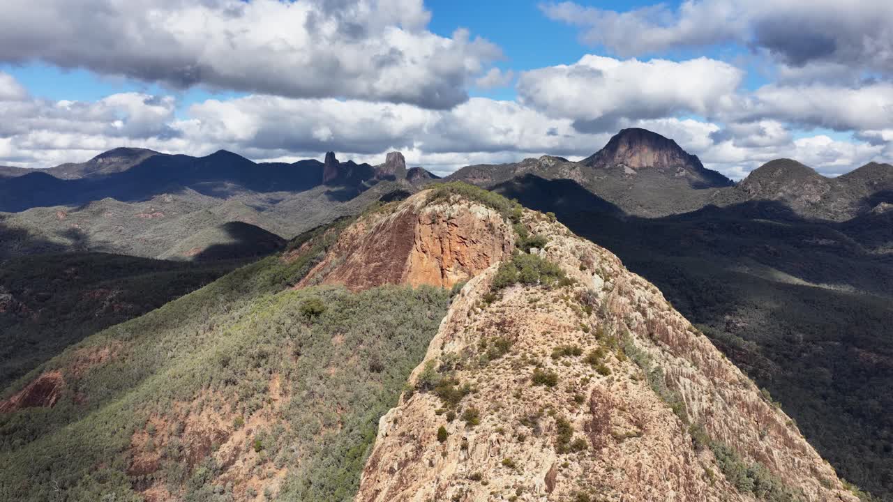 Drone camera glides above rugged Split Rock ridge, revealing expansive mountain landscape under dynamic daylight and scattered clouds in Coonabarabran, New South Wales