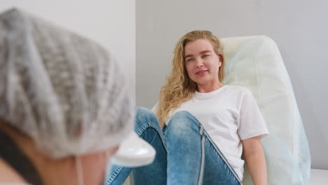 client in white shirt and jeans smiling and relaxing on chair during pedicure session while nail technician slightly visible in foreground, showing comfortable and friendly salon experience