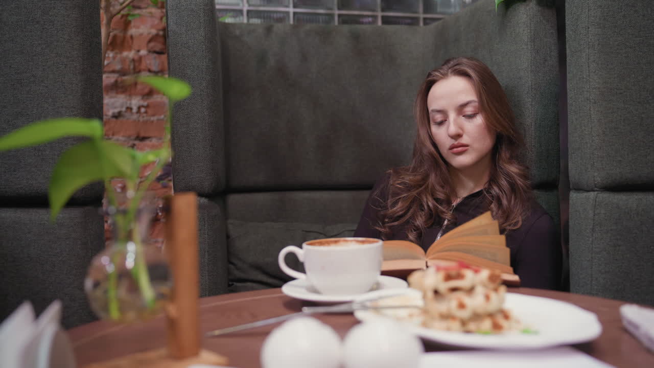 Lady dressed in black sits inside cozy booth, leaning gently on cushion while reading brown book and flipping page, surrounded by cup of latte, waffle plate, decorative plant, and soft natural light