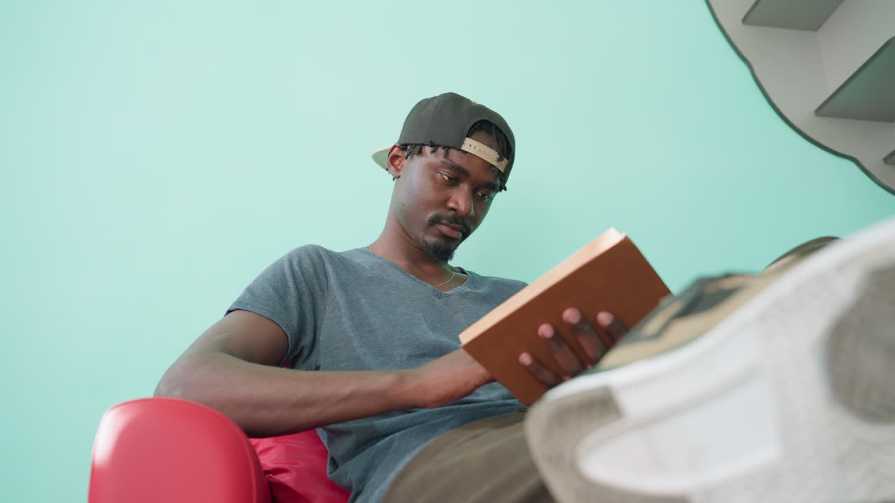 Relaxed man in cap sits on red couch with crossed leg, holds open book, reads quietly, flips pages slowly, sneaker foreground and turquoise wall behind create cozy study mood