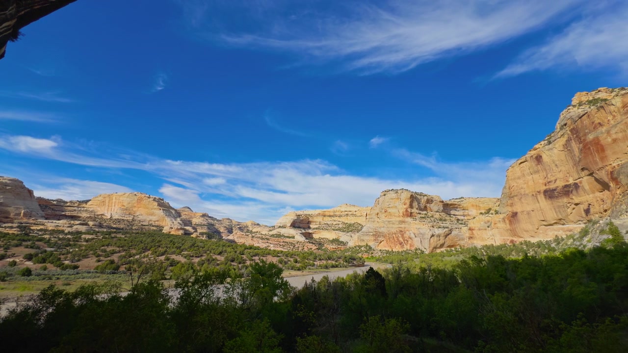 Cielo azzurro brillante sopra un vasto paesaggio di canyon con scogliere e vegetazione verde, un punto panoramico che risplende della luce del pomeriggio.