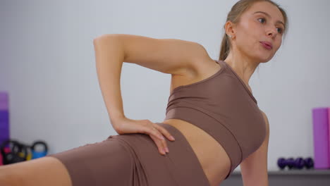 mujer con ropa deportiva marrón coloca la mano en la cintura mientras se estira y conversa, el fondo presenta equipos de gimnasia borrosos, incluidos pesos y esteras de ejercicio