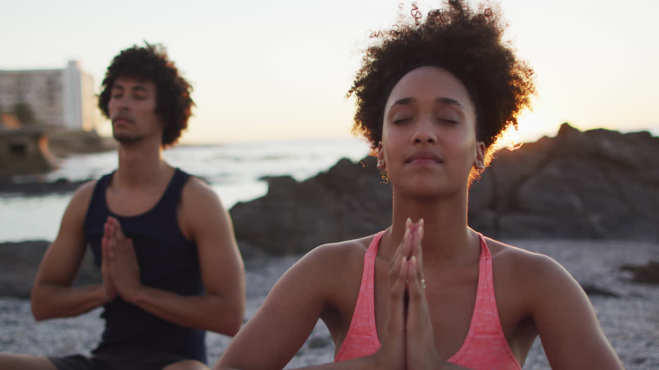 Coppia afroamericana che pratica yoga e medita insieme sulle rocce vicino al mare al tramonto