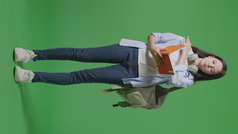 full body of tired asian teen girl student with a backpack reading book while standing in the green screen background studio