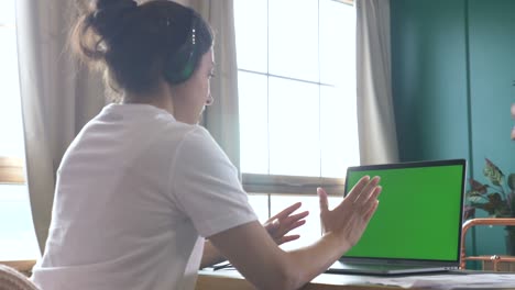 vista trasera de una joven india que trabaja en una conferencia en línea, entrenando con personas, usando una computadora portátil con una pantalla verde. habitación brillante en casa