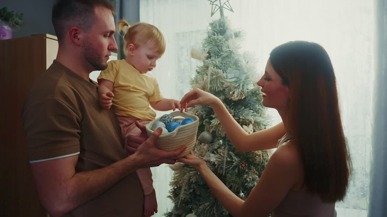 Pais amorosos pendurando cuidadosamente enfeites festivos enquanto interagem com sua filha bebê, criando memórias natalinas emocionantes dentro de uma sala de estar aconchegante durante a época de Natal