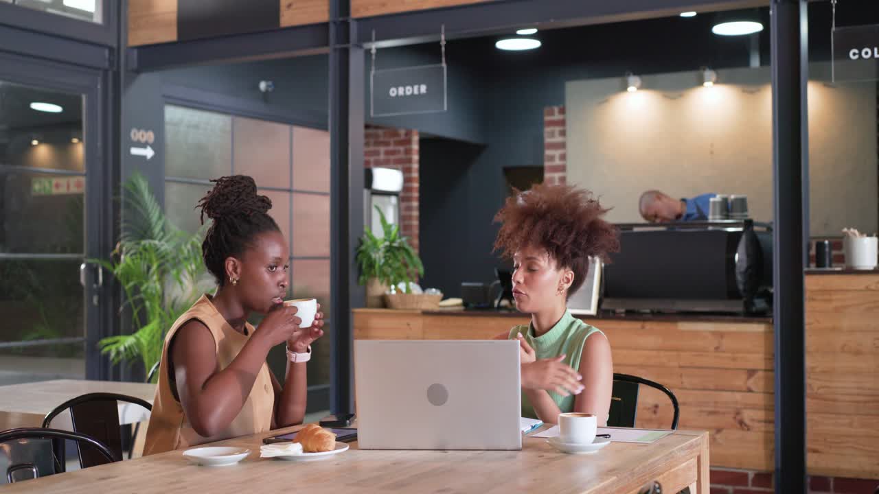 Two African American women working on silver laptop at wooden cafe table after green top gesturing