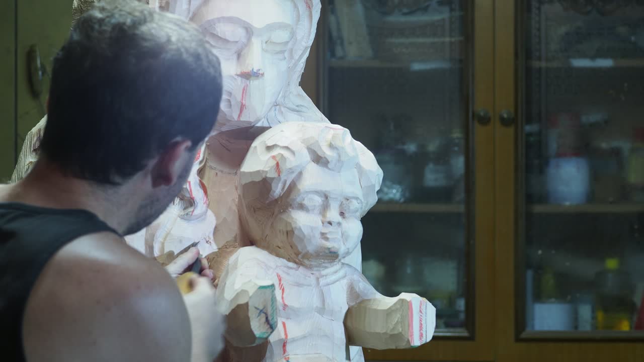 Craftsman concentrates while carefully carving the wooden figure of baby Jesus on large Madonna sculpture, working in a traditional studio surrounded by tools and wood-finishing materials, real time
