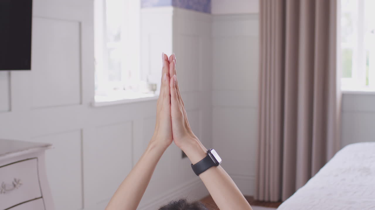 Close up of woman's hands coming together in yoga pose before camera tilts down to show her sitting on exercise mat in bedroom - shot in slow motion