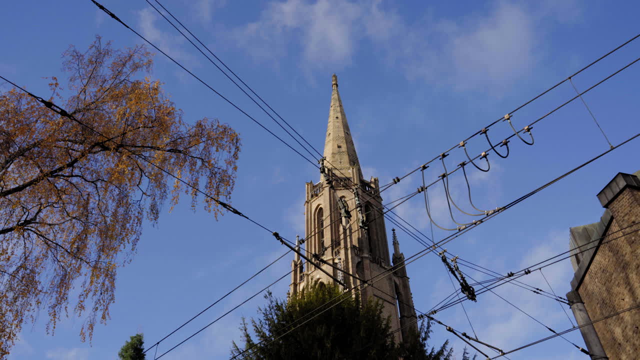 른의 고 양식의 교회 첨탑은 고 푸른 하늘을 배경으로 아 있으며, 황금색 가을 잎과 교차하는 상공 전선으로 여 있으며, 역사적인 건축과 도시 요소를 생생한 장면으로 혼합합니다.