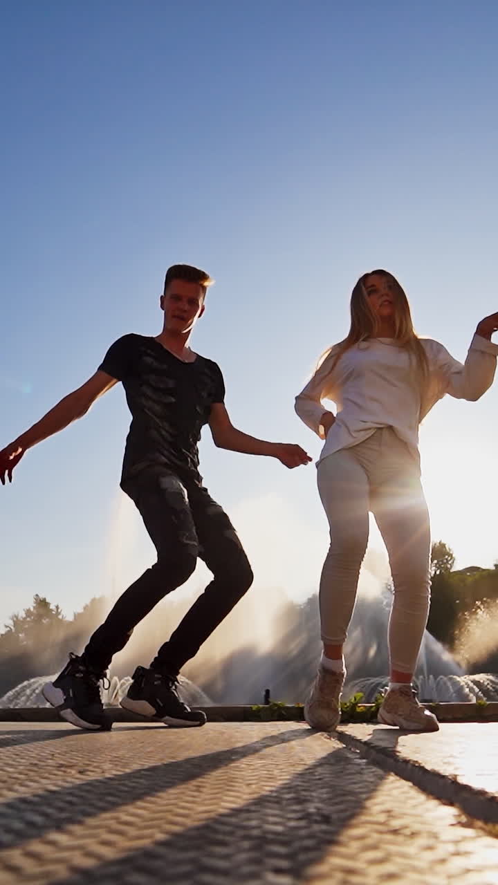Dancers against beautiful fountain. Young couple performing a modern dance on the urban background. Contemporary freestyle dance of teens outdoors through the leaks of sun. Vertical video