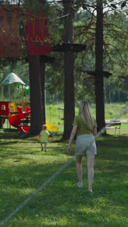 long haired woman in shorts follows little son walking to large colorful playground on glade in old pine forest backside view slow motion