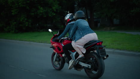 two sisters ride a red power bike through a street with helmets on, passing parked cars and a person walking nearby, as they drive into a shaded path with trees and buildings around