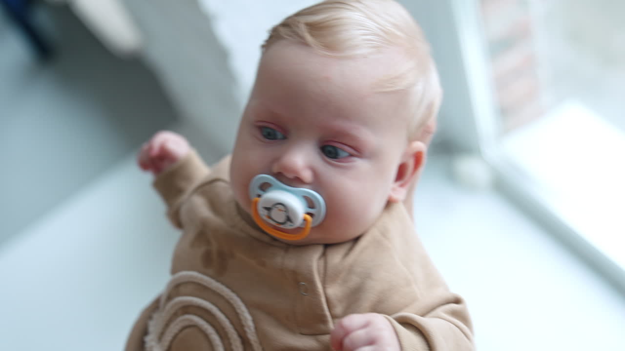 Cute blond baby boy with big blue eyes is waved in hands. Kid sucking a pacifier and looks at mom attentively.