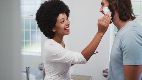 mujer afroamericana aplicando crema de afeitar en la cara de su marido en casa