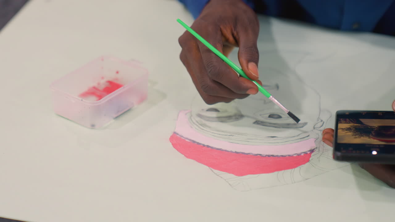 Black artist painting portrait with pink highlights, closeup hand applying watercolor on canvas, phone reference showing face, plastic paint tray, demonstrative tutorial mood, focused technique