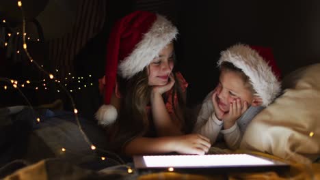 happy caucasian siblings wearing santa hats lying in makeshift tent, using tablet