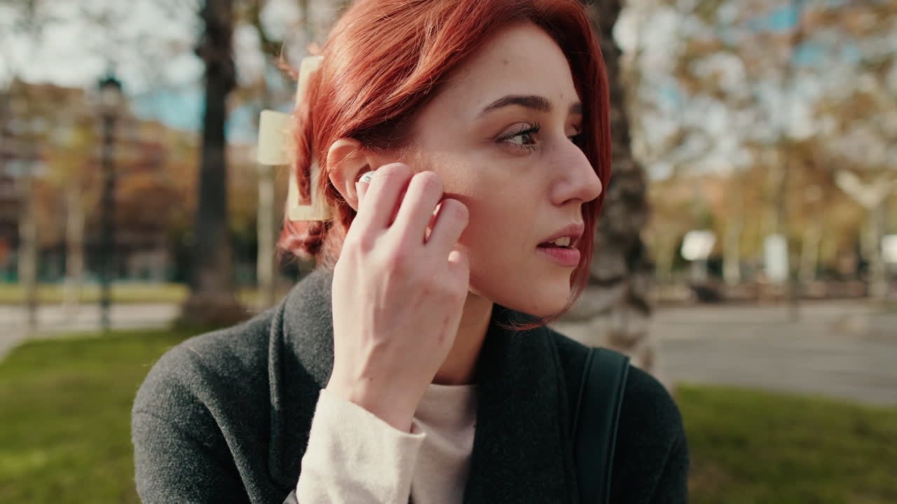 Young Woman with Red Hair Listening to Music with Wireless Earbuds in Park