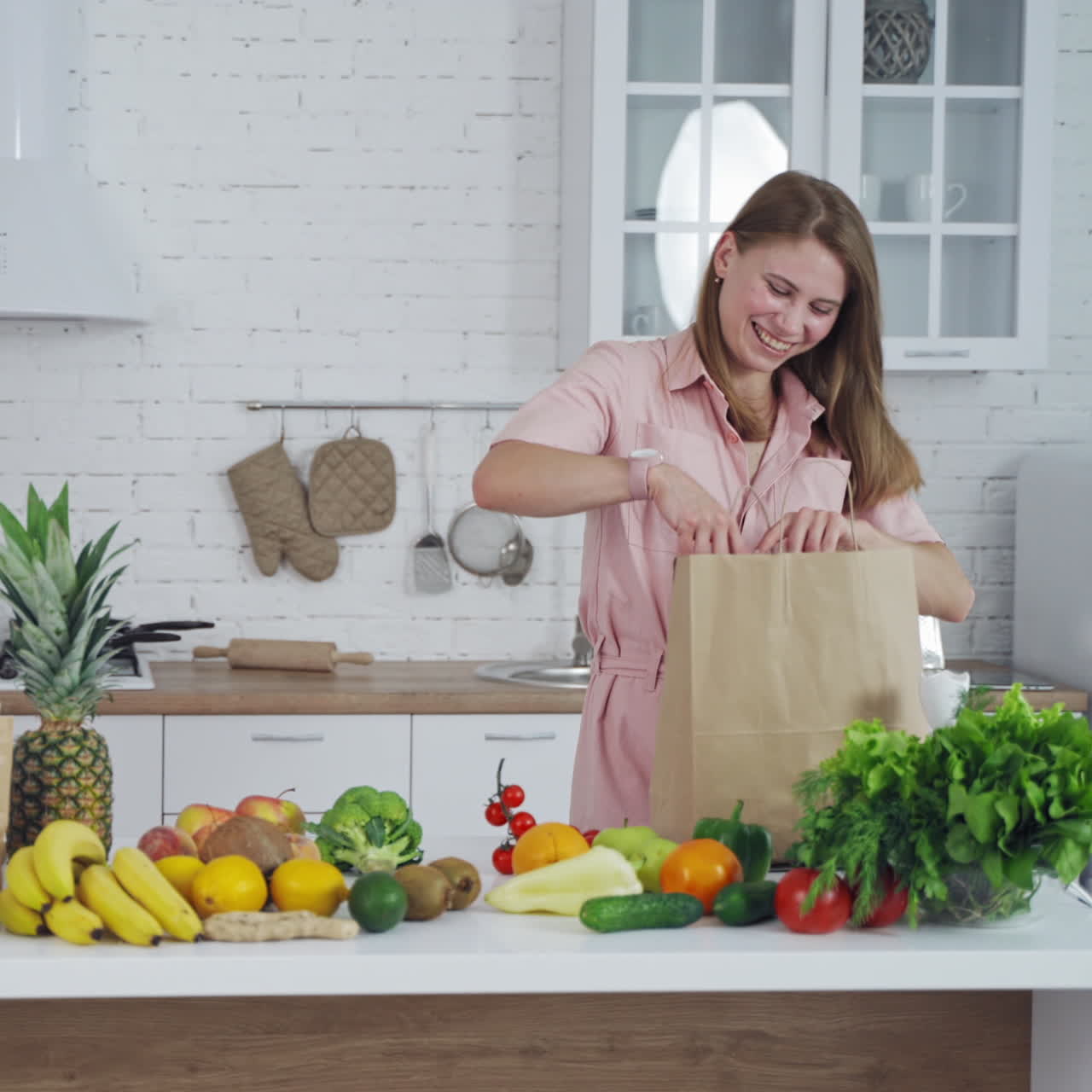Pretty woman taking out fresh fruit from paper shopping bag at home. Young female holds an orange in the light kitchen. Healthy fruit and vegetables on the table.