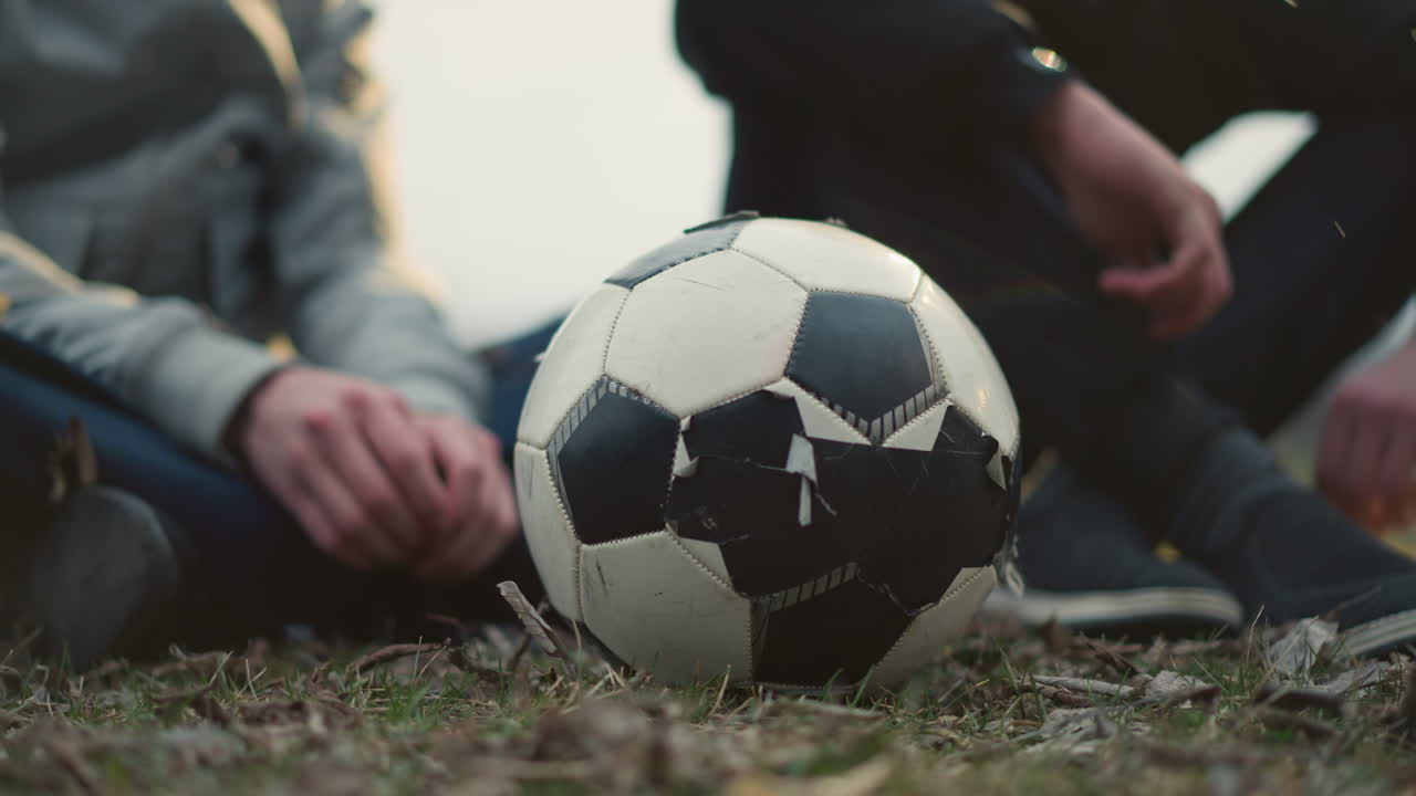 Close-up of a worn soccer ball on a grassy field with two people seated behind it, one of them in a black outfit appears to be picking something from the ground