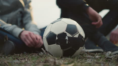 close-up of a worn soccer ball on a grassy field with two people seated behind it, one of them in a black outfit appears to be picking something from the ground