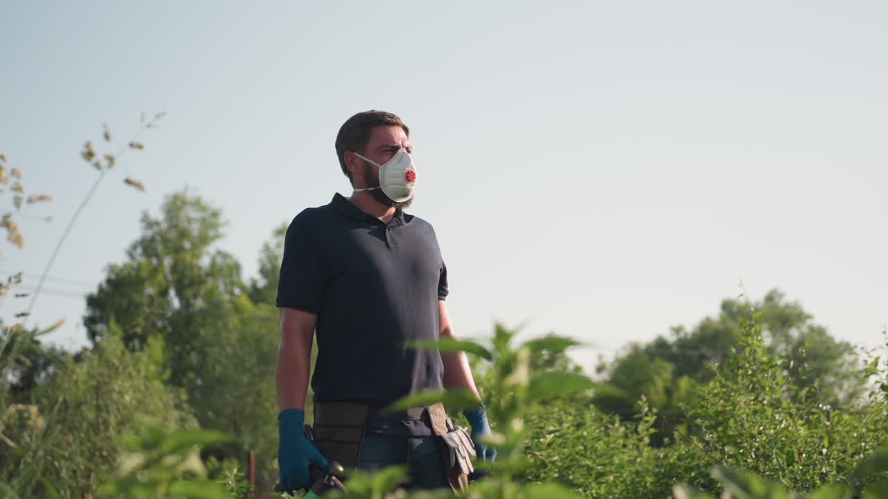 Side view of gardener wearing protective mask and blue gloves wiping sweat from forehead while holding green spray bottle, surrounded by tall grass and dense greenery