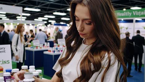 female professional browsing nutritional supplement line, standing near display table at wellness industry expo, examining product bottles with focused interest