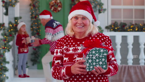 Portrait-of-grandmother-woman-presenting-gift-box-smiling-near-decorated-Christmas-house-with-family