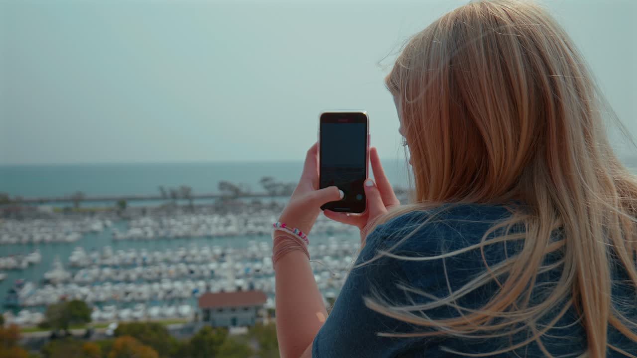 una adolescente rubia caucásica tomando fotografías con su teléfono celular de un puerto en california