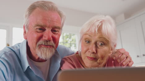 retired senior couple in kitchen at home making video call using laptop