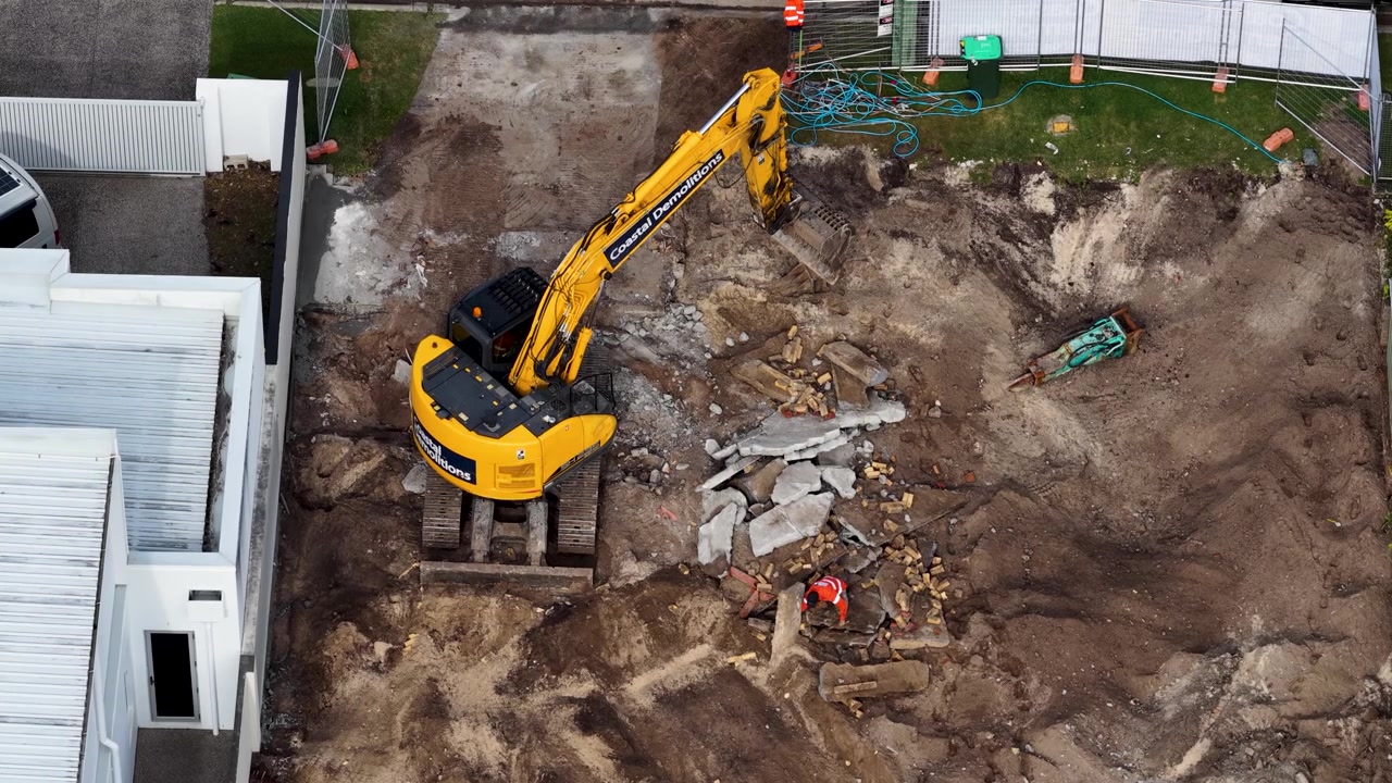 Højt vinkelbillede af tunge maskiner, der rydder betonmurbrokker og jord på en nedrivningsplads for boliger.