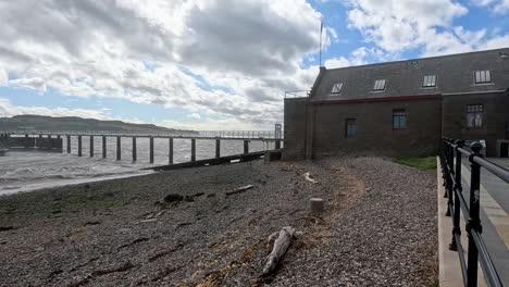 wide shot pans along a pebbled shoreline, revealing a pier, bridge, and waterside building under dramatic clouds in broughty ferry, scotland