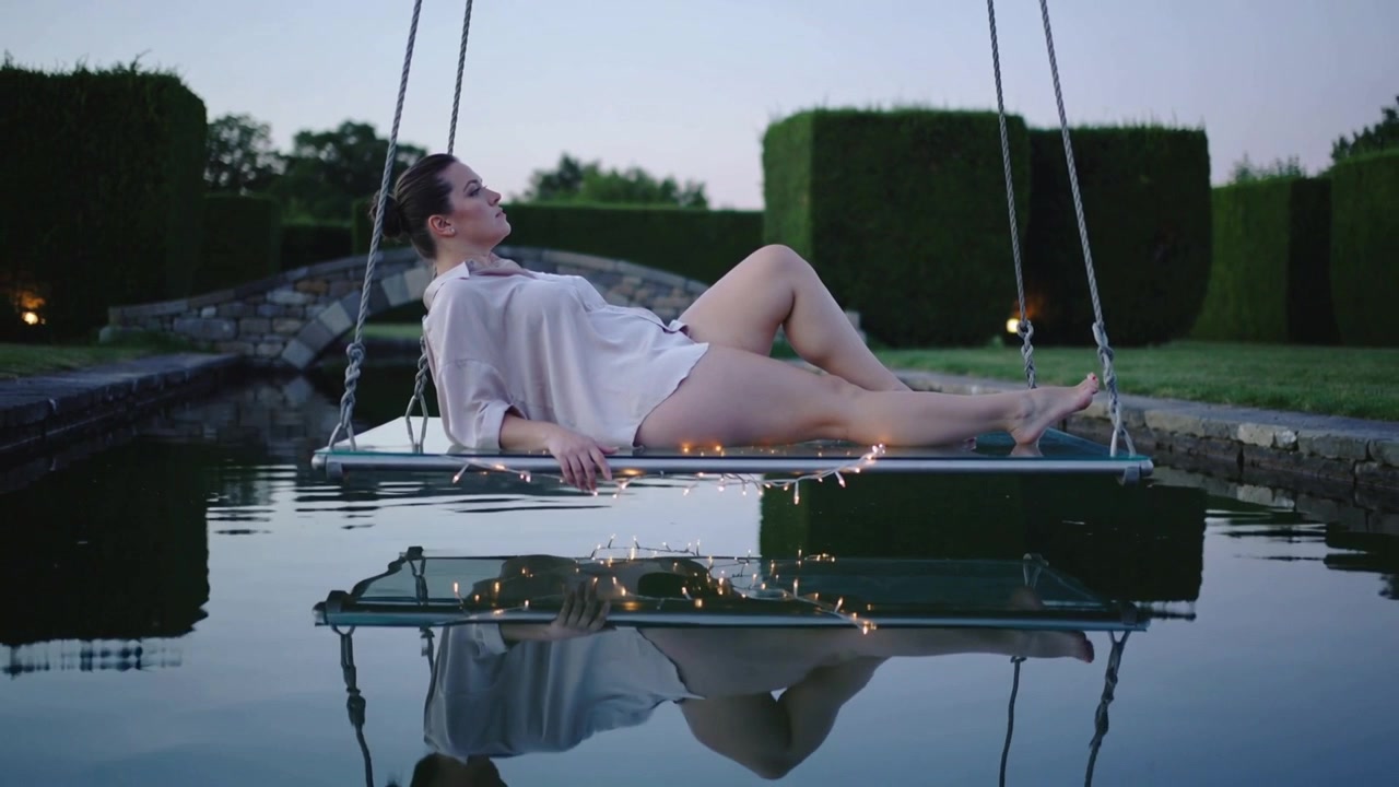 Modella sdraiata in camicia che aggiusta la posa su un'altalena sopra una piscina mentre il crepuscolo si intensifica, con luci fatate che si riflettono.
