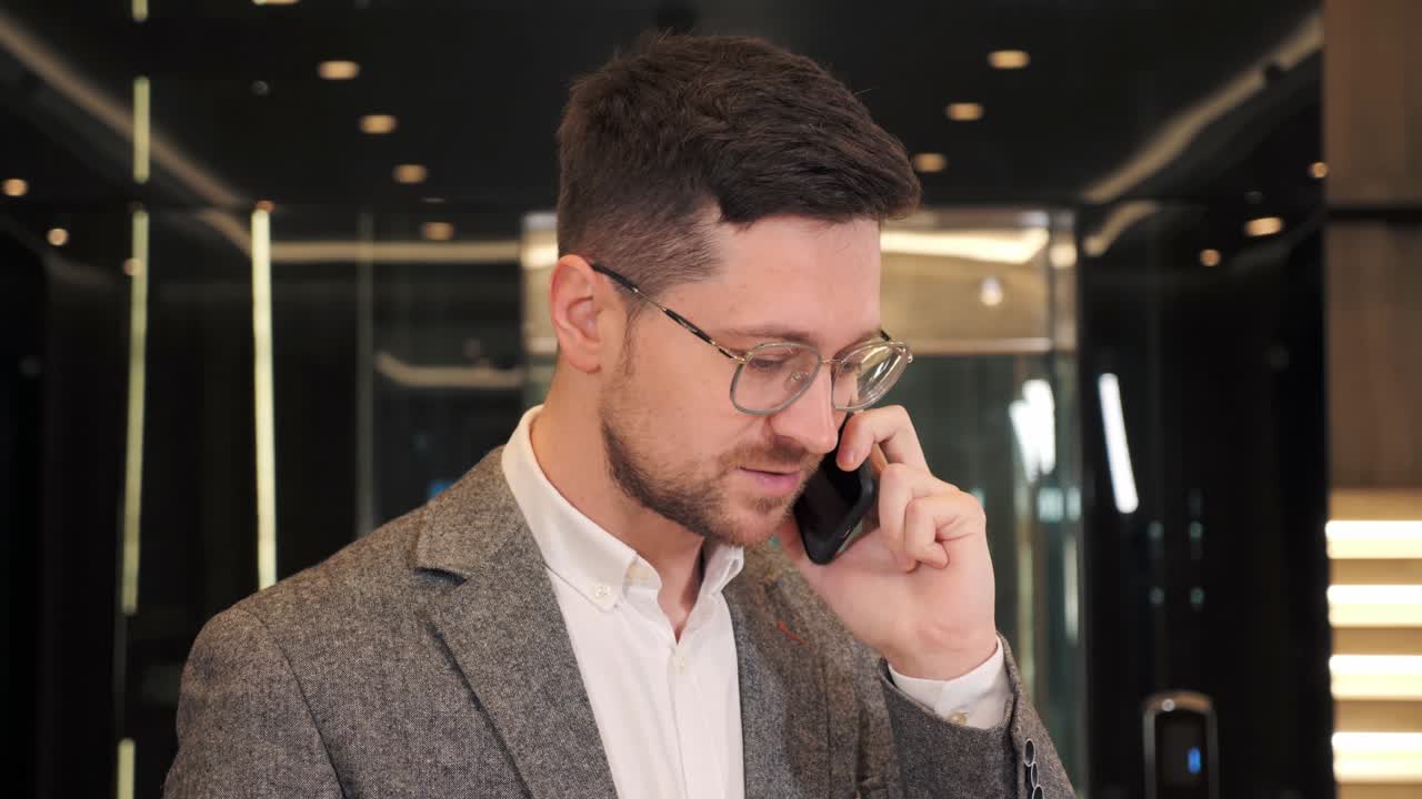 Closeup smiling bearded man in glasses talking on mobile phone in office hall. Close up of cheerful businessman having phone call near elevator. Portrait of happy business man calling phone indoor.