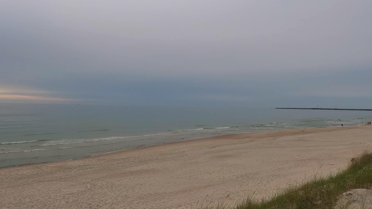 Calm Baltic Sea waves wash onto the sandy shore of Smiltynė under a moody overcast sky.