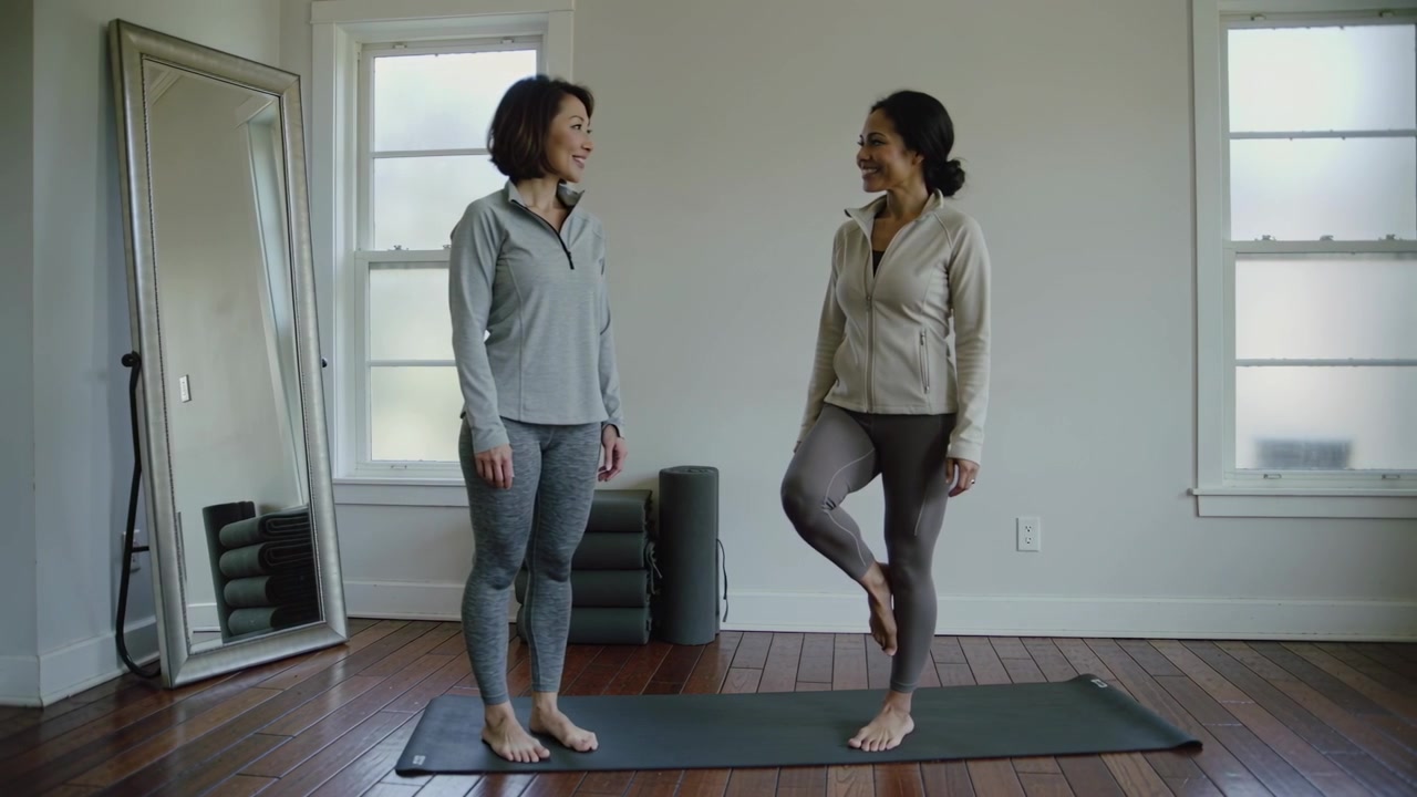 Balancing right woman lifting left knee after smiling, practicing balance on yoga mat in leggings