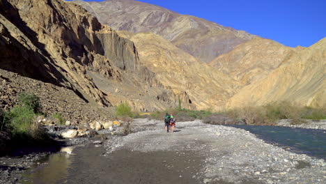 tilt down shot to a group of hikers walking at the river bank with mountains in the background on a warm, sunny day