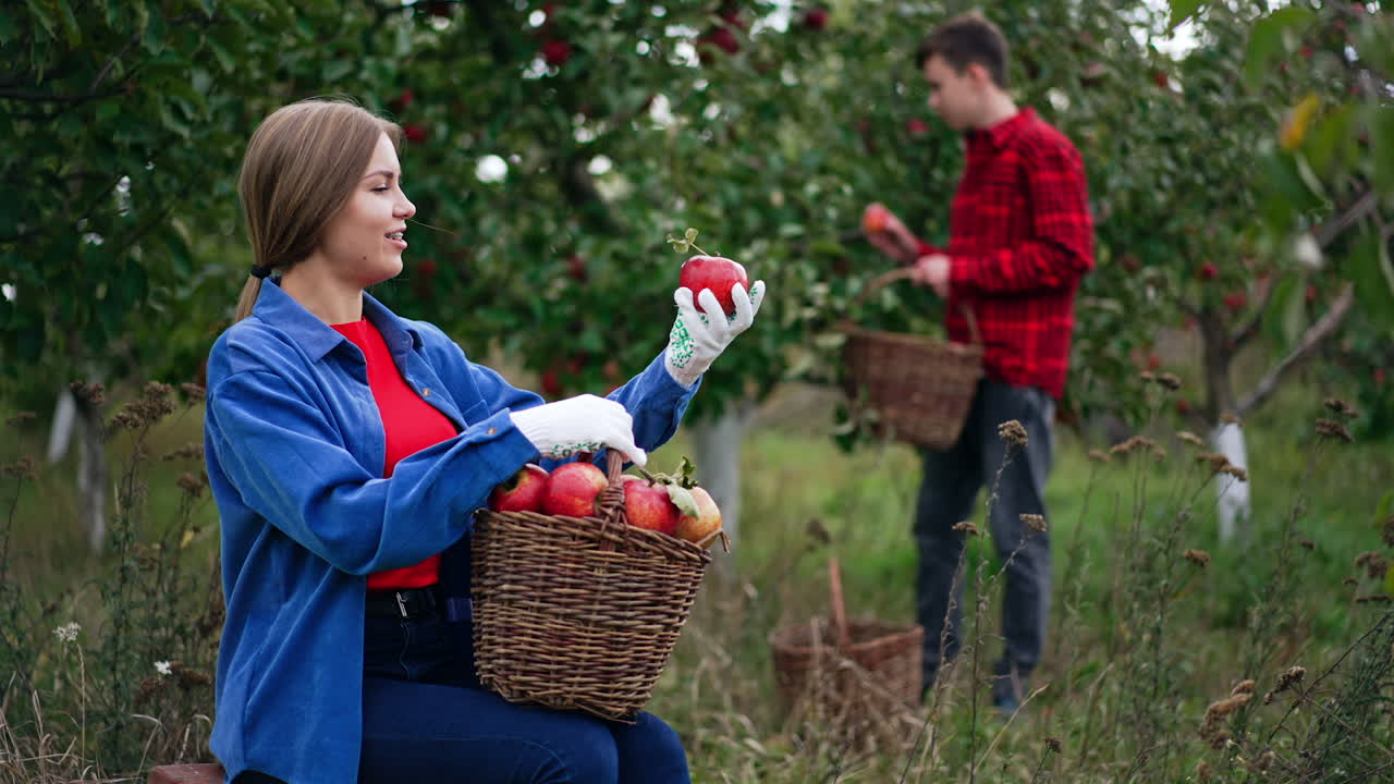 Long-haired lady sitting in the garden enjoying the harvest she's picked. Woman takes apple from a basket on her laps and shows it to the boy at backdrop.