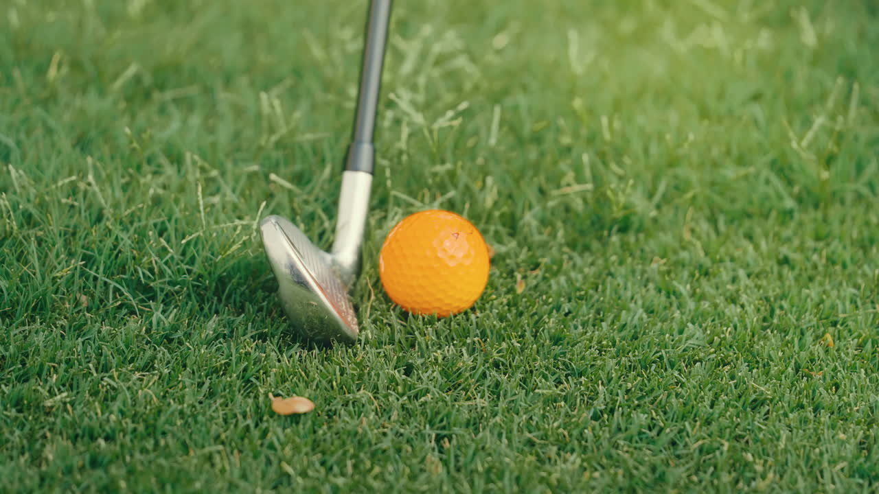Slow-motion clip of an iron striking an orange golf ball sitting near the edge of the fairway. Grass and debris fly as the swing comes through, showing dynamic energy and technical precision.