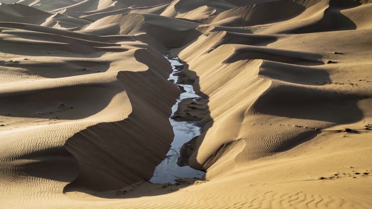 Drone che filma scivolando lungo un avvallamento tra le dune, mostrando i cambiamenti di luce su uno stretto canale d'acqua.