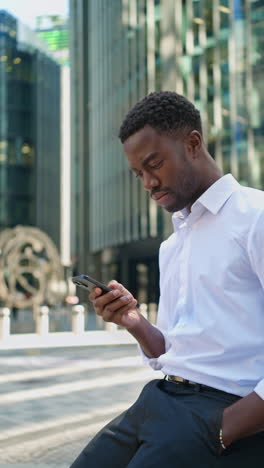 vertical video of young businessman in shirt sleeves using mobile phone standing outside offices in the financial district of the city of london uk shot in real time 1